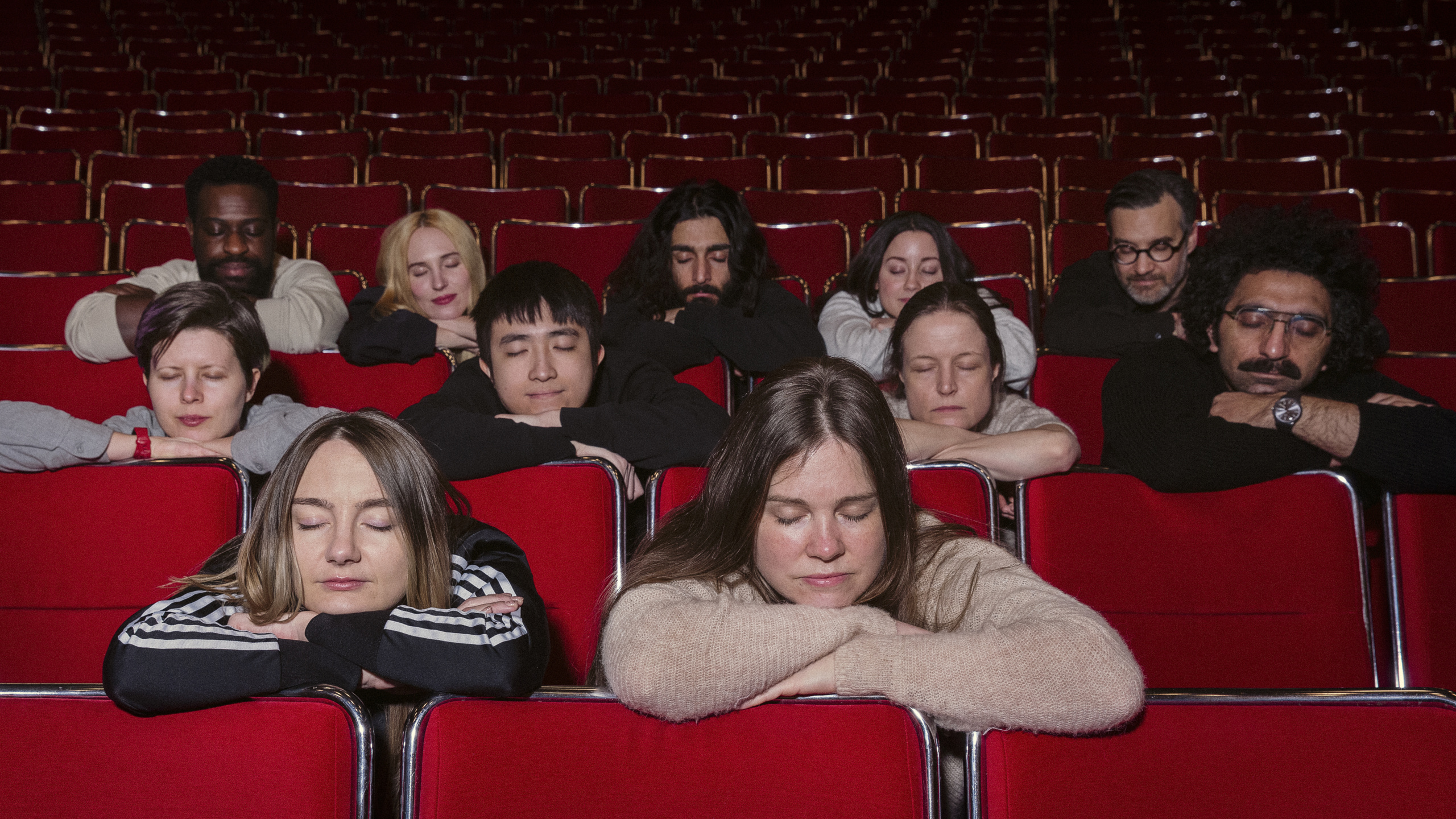 Students and teachers are sitting and leaning on the seats in front of them with their eyes closed in a cinema.
