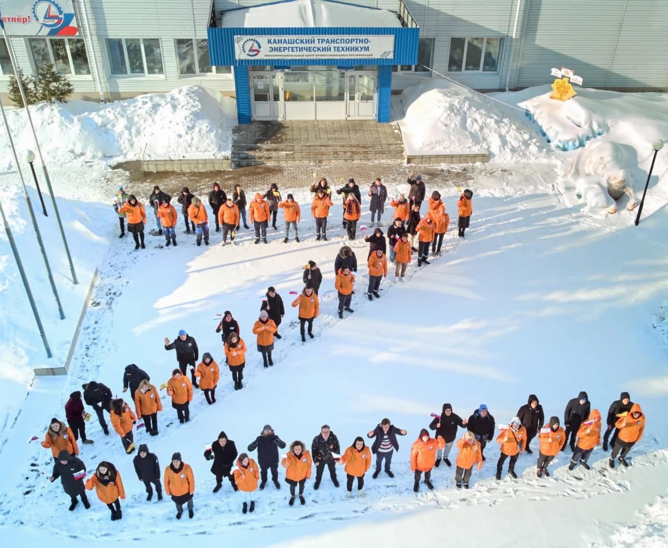 A snowy school frontyard pictured from above. A group of people wearing orange and black clothes stand in a formation in the shape of the letter Z, and wave Russian flags. 
