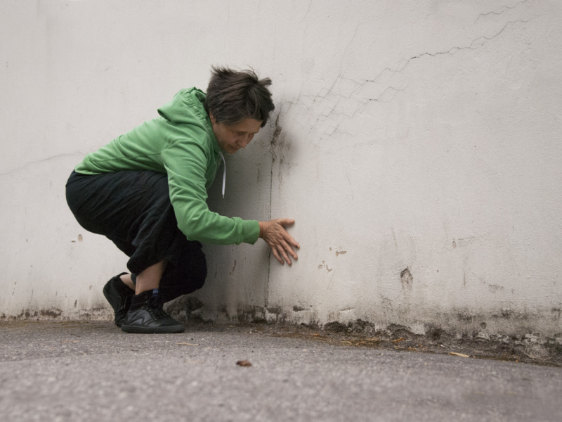 Henkilö vihreässä hupparissa on kyyryssä ulkona asfaltilla ja koskettaa käsillään vaaleaa betoniseinää.