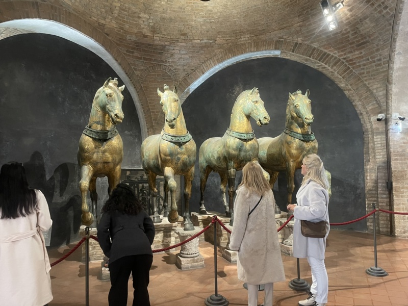 Neljä pronssista hevospatsasta rintamasuunta eteenpäin tiilisessä sisätilassa, jonka taustalla kaksi holvikaarta. Etualalla neljä museovierailijaa katsoo hevosia punaisen nauhan takaa.